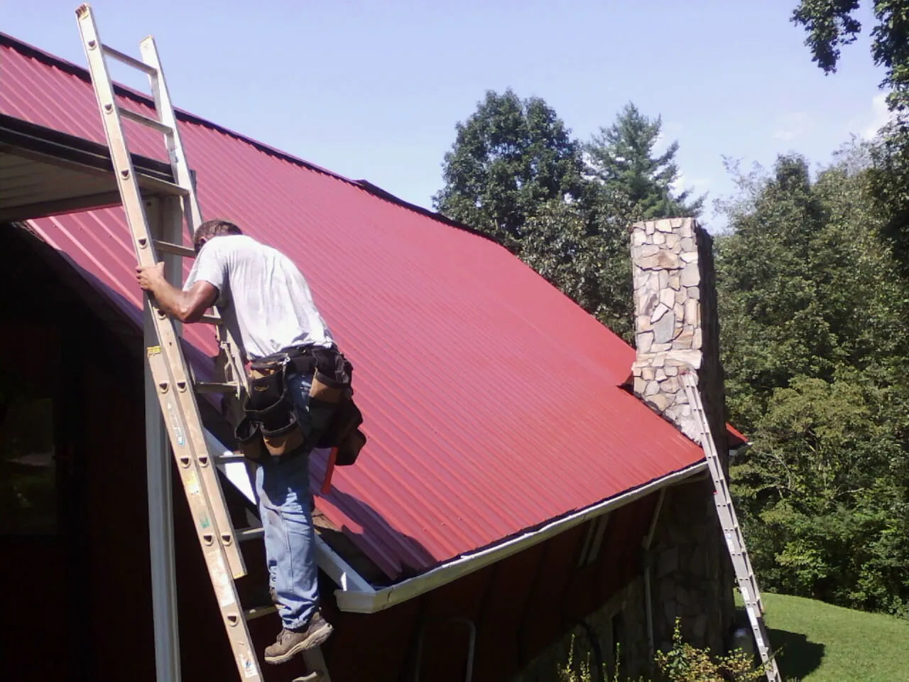 Roofer on a ladder beside a red metal roof and stone chimney — Soffit & Fascia Repair by Roofing Brothers in Chesterton